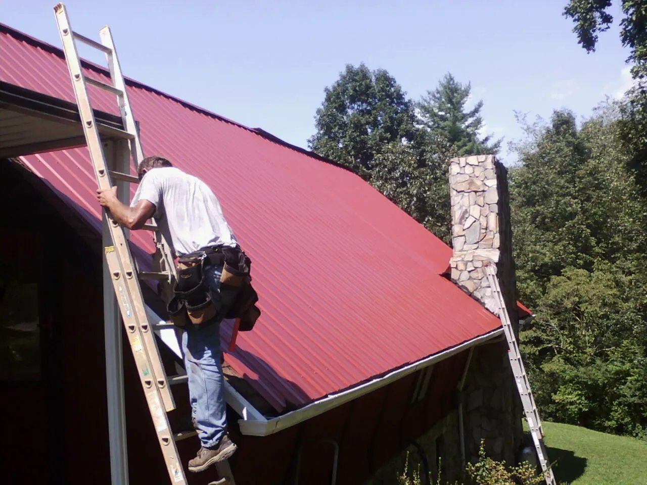 Roofer on a ladder beside a red metal roof and stone chimney — Soffit & Fascia Repair by Roofing Brothers in Naturita